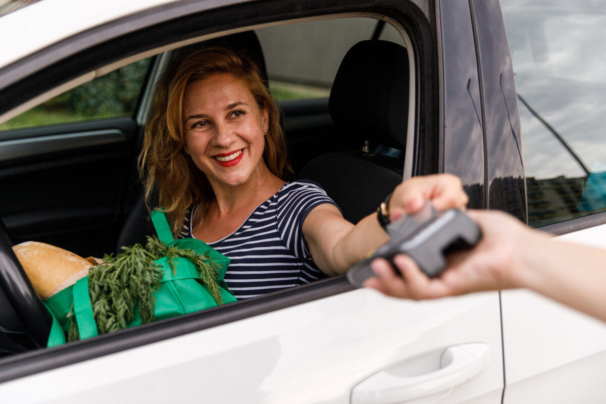 Selective focus shot of cheerful mid adult woman sitting in her car, in a drivers seat and playing for groceries with a credit card during a curbside pickup.