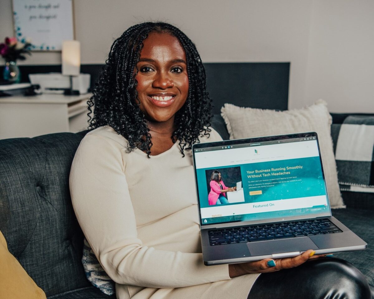 Ruth Aliu, founder of Techity Consulting, smiles while sitting on a couch and holding a laptop displaying her business website, techityconsult.ca. The screen reads “Your Business Running Smoothly Without Tech Headaches,” reflecting her mission to help Canadian small businesses manage their technology. The setting is cozy and professional, hinting at the approachable, tailored tech support she offers through her .CA website.