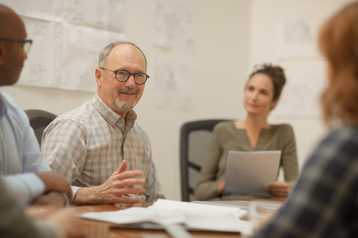 Senior man listens to coworker in meeting with work colleagues while sitting at table in design office
